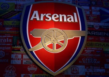 Logo Arsenal Football Club berwarna merah, emas, dan biru tua yang menyala, menampilkan meriam emas mengarah ke timur, dipasang di dinding yang ditutupi bendera dan banner pendukung Arsenal dari seluruh dunia.