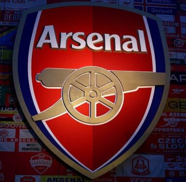 Logo Arsenal Football Club berwarna merah, emas, dan biru tua yang menyala, menampilkan meriam emas mengarah ke timur, dipasang di dinding yang ditutupi bendera dan banner pendukung Arsenal dari seluruh dunia.