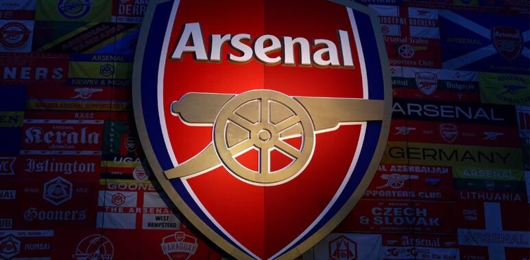 Logo Arsenal Football Club berwarna merah, emas, dan biru tua yang menyala, menampilkan meriam emas mengarah ke timur, dipasang di dinding yang ditutupi bendera dan banner pendukung Arsenal dari seluruh dunia.